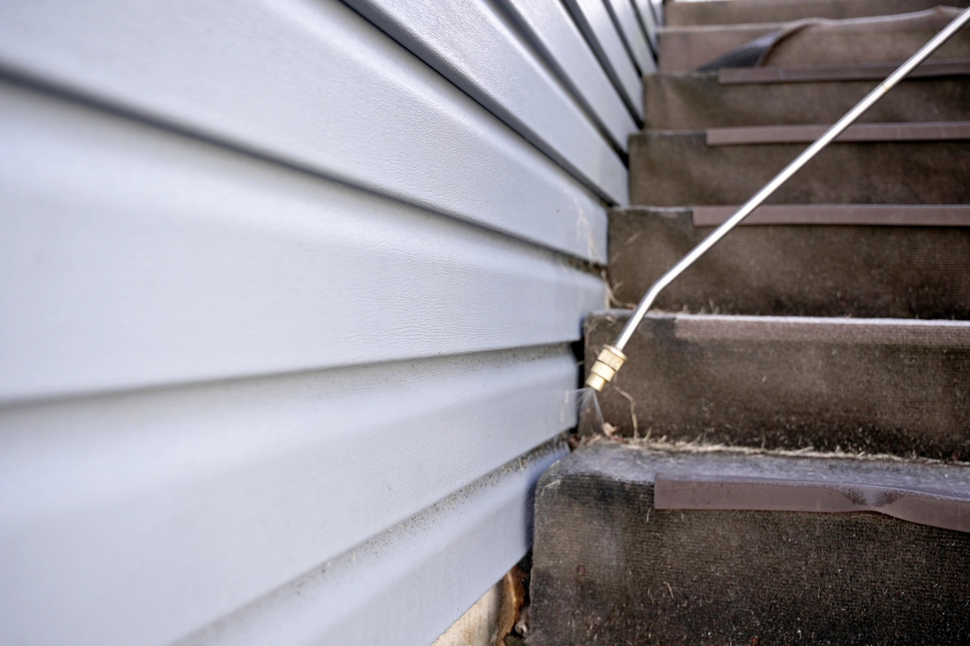 Spray lance applying a surface treatment along the base of a home's weatherboard cladding beside external steps